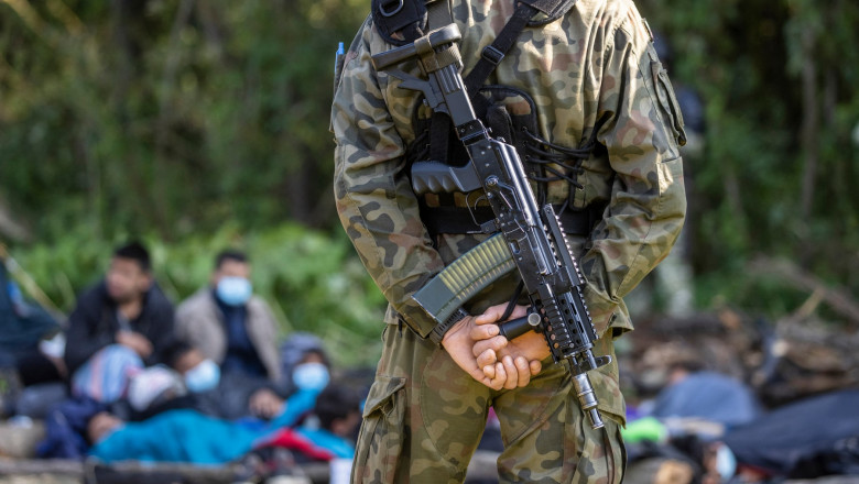 un soldat polonez cu arma în spate păzește migranți afgani de la granița cu Belarusul