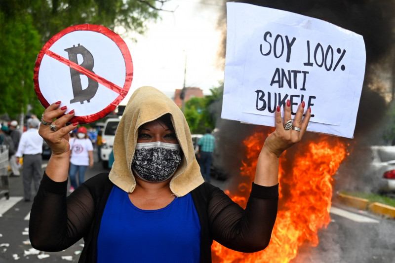 Femeie cu văl pe cap și bannere anti-bitcoin, în spate arde un cauciuc