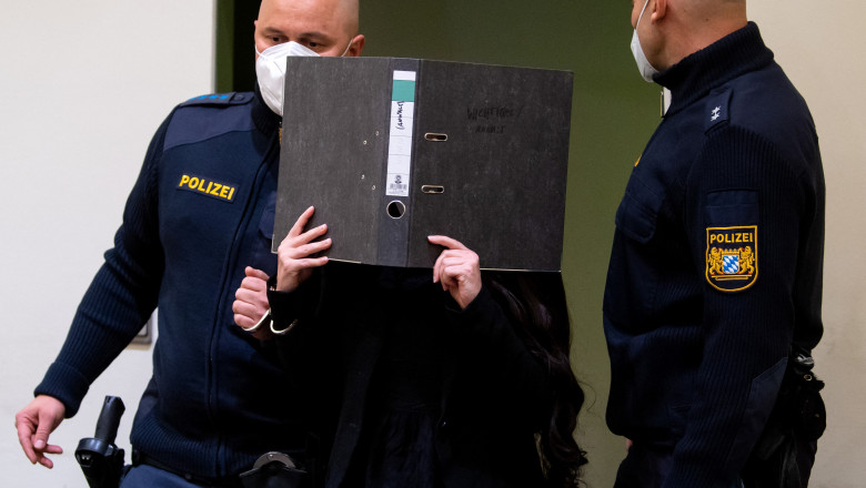 femeie își acoperă fața cu un dosar, flancată de polițiști