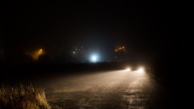 car headlight cones in night fog at field behind city