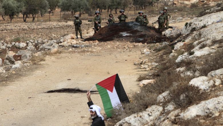 Un bătrân flutură un steag palestinian în fața unor militari israelieni.