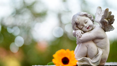 statuie a unui inger pazitor care doarme langa care este pusa o floare, pe un mormant