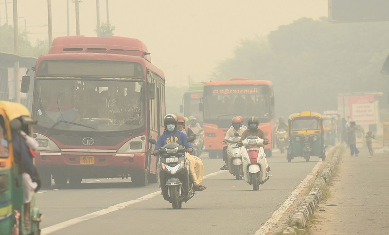 Stradă din New Delhi cu motocicliști și autobuze, mergând prin fumul produs de poluare