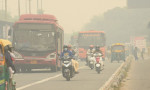 Stradă din New Delhi cu motocicliști și autobuze, mergând prin fumul produs de poluare