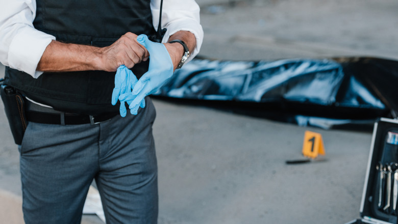 cropped image of policeman with gun in holster putting on latex gloves at crime scene with corpse in body bag