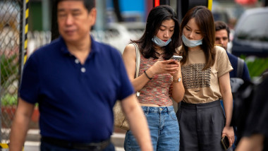 două femei cu mască se uită pe un telefon la o coadă de testare Covid, în timp ce un bărbat îmbrăcat într-un tricou albastru se află în fața lor