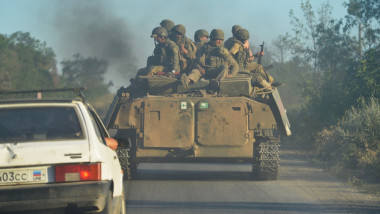 militari urcați pe un tanc în mișcare și o mașină în urma lor