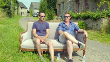 doi barbati stau pe o canapea pusă pe strada dintr-o localitate cu cladiri vechi