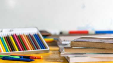 Coloring pencils, pens and closed books and notebooks on top of a yellow wooden table, with a white school board in the background