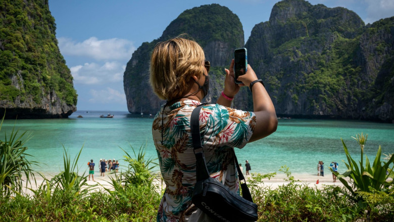 Femeie cu mască face o fotografie cu telefonul mobil unei plaje tropicale