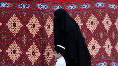 femeie cu burka si copil de mana, covor rosu pe fundal