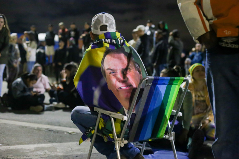 protestatar stă pe un scaun pliant cu steag pe spate care are chipul lui Bolsonaro