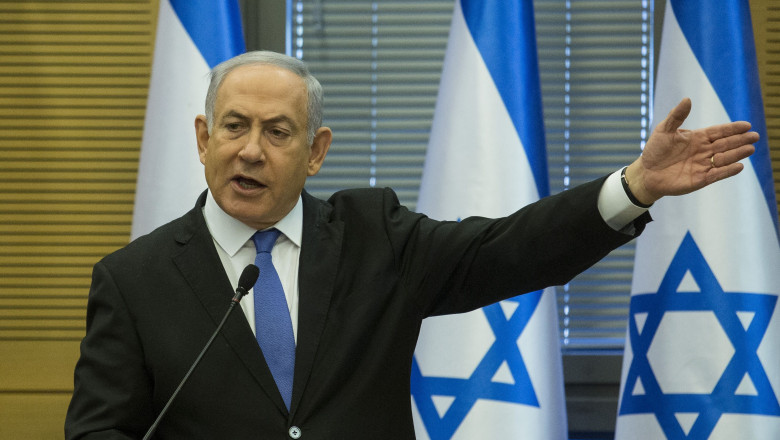 Benjamin Netanyahu cu steaguri israeliene în spate gesticulează cu mâna stângă