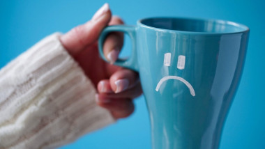 O mână de femeie ţine o ceaşcă de cafea pe care este desenata o faţă tristă.