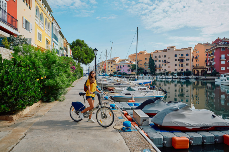 Smiling woman tourist with bicycle on pier with yachts and boats at beautiful harbor of Port Saplaya near Valencia with colorful houses