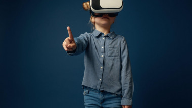 Discoverer of future. Little girl or child pointing to the empty space with virtual reality glasses isolated on blue studio background. Concept of cutting edge technology, video games, innovation.
