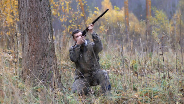 Un vânător trage în sus cu o puşcă în pădure, pe timp de toamnă.