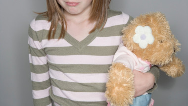 O fetiţă cu faţa parţial acoperită ţine în braţe un urs de jucărie.