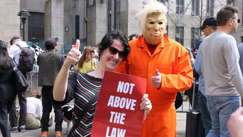 Protestatară anti-Trump la New York cu un banner inscripționat "not above the law" și o persoană îmbrăcată într-o uniformă portocalie de închisoare cu mască de trump