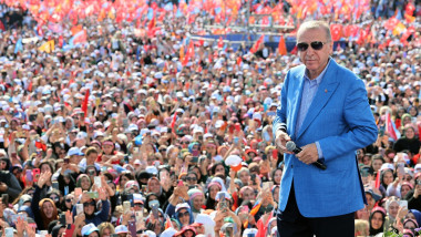 erdogan in sacou si ochelari de spare pe scena lșa miting politic cu multi oameni in spate