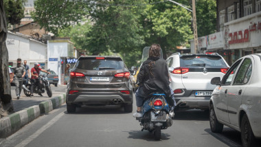 woman sits on the back seat of a moped in Iran with her hair uncovered; the license plate is not visible