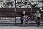 Femeie în vârstă stă pe bancă, pe fundal fiind o clădire distrusă