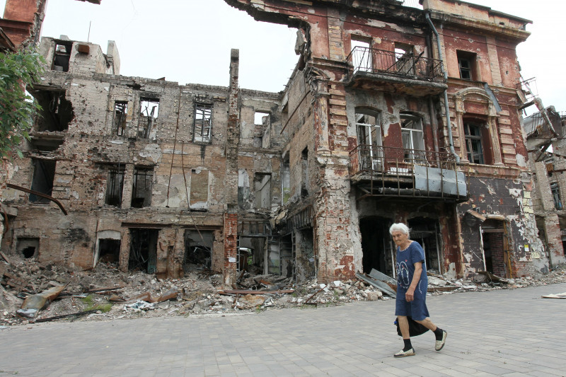 Femeie în vârstă merge prin fața unei clădiri în ruine