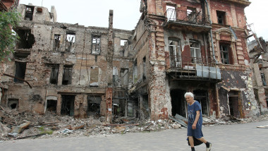 Femeie în vârstă merge prin fața unei clădiri în ruine