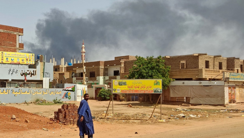 Fum în aer deasupra unui oraș din Sudan în timp ce un trecător în albastru merge pe stradă
