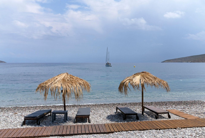 Straw beach umbrellas and bedchairs look out over idyllic beach scene with blue sky, Livadia village, Tilos island, Dodcanese, Greece