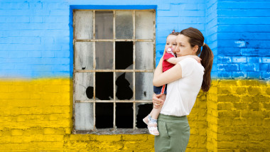 o femeie tine strans in brate un copil mic care are o suzeta in gura, in fata unui zid cu steagul ucrainei si un geam spart