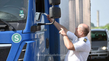 un politist de frontieră, bărbat, îi întinde niște acte unui șofer aflat într-un camion