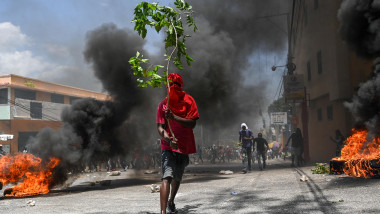 un barbat care tine o ramura de copac pe o strada din Haiti, se vad cauciucuri care ard pe srada