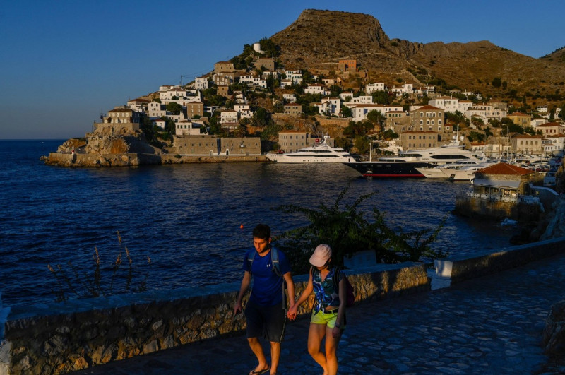 Hydra, Greece. 24th July, 2020. A couple walks hand in hand past the harbour. The island of Hydra, which covers almost 50 square kilometres, is 65 kilometres south-west of Athens. Cars, neon signs and plastic chairs are prohibited on the island. Credit: A