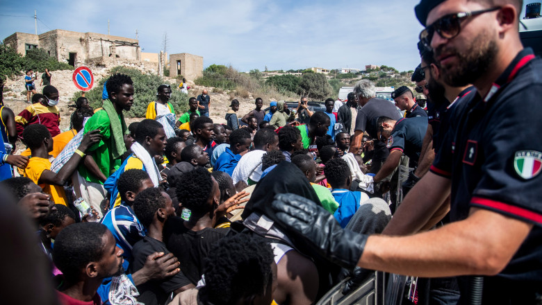 Migranți la coadă în Lampedusa, în timp ce un polițist italian se uită la ei