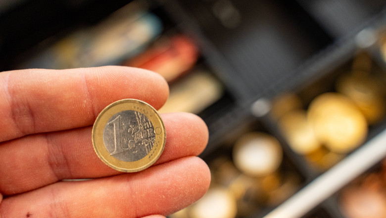 Bavaria, Germany - 2 September 2023: Hand holding a 1 euro coin in front of a cash register with cash *** Hand hält eine