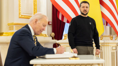 Joe Biden semnează la un birou, privit de Volodimir Zelenski