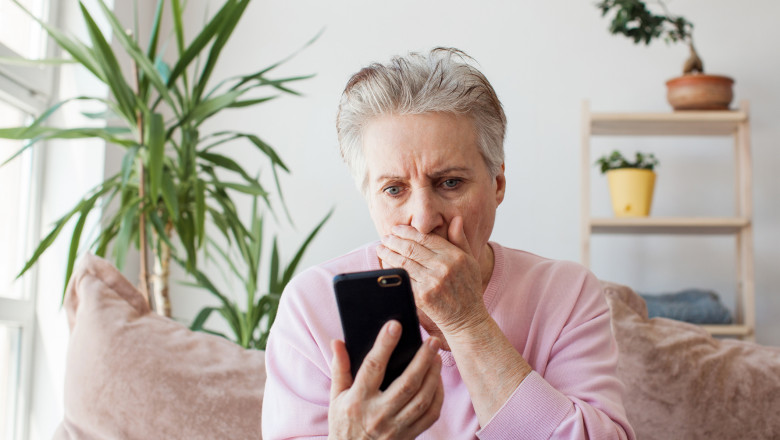 o femeie batrana cu un telefon in mana si cealalta mana la gura