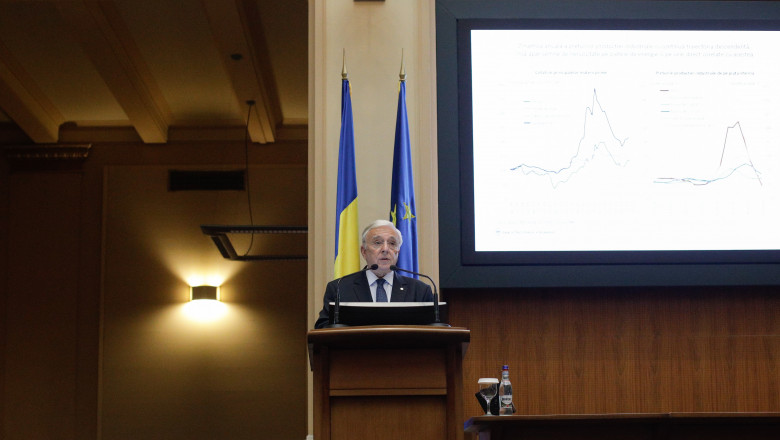 mugur isărescu prezintă un raport de la un pupitru, cu steaguri în spate și un ecran in dreapta