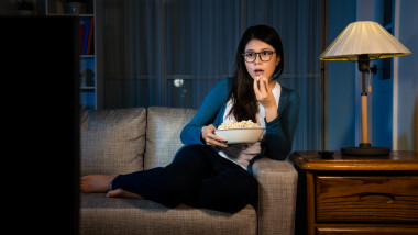 o femeie cu ochelari sta pe canapea, mananca popcorn si se uita la tv