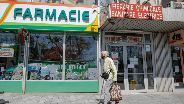 o femeie in varsta trece pe trotuar in fata unei farmacii si a unui magazin