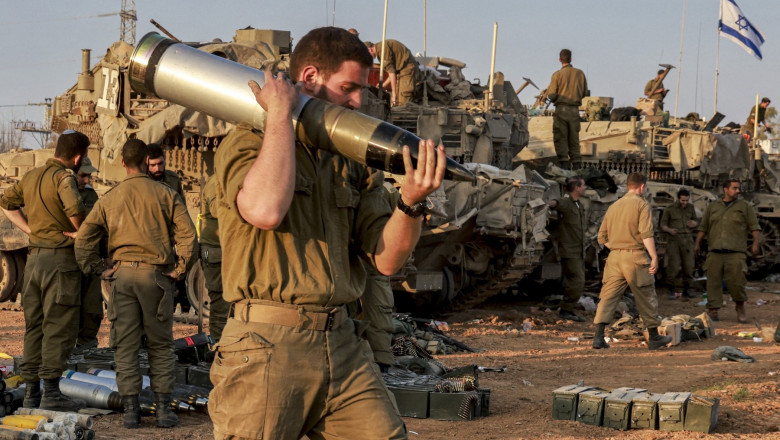 An Israeli soldier carries a heavy shell past battle tanks deployed at a position along the border with the Gaza Strip