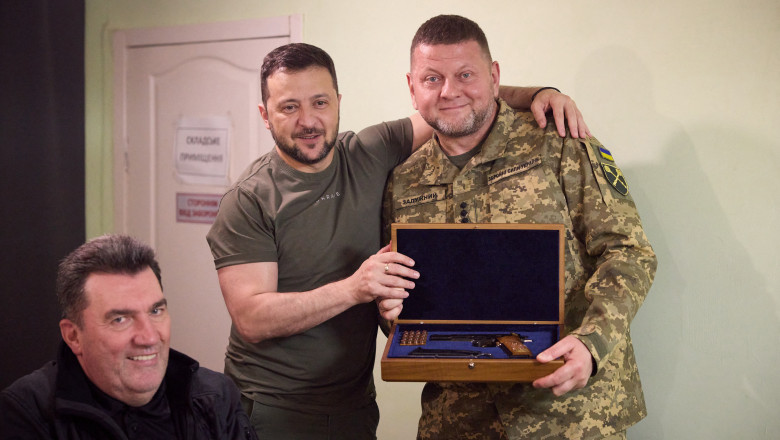 Volodimir Zelenski îl îmbrățișează pe Valeri Zalujnîi care ține în brațe un pistol în cutie, făcut cadou