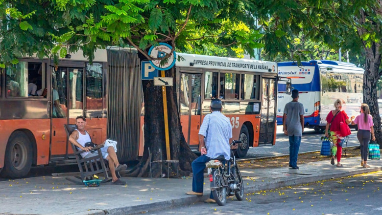A Cuban person sits in a rocking chair by a bus stop speaking with a man in a cycle. Other people walking in the area.