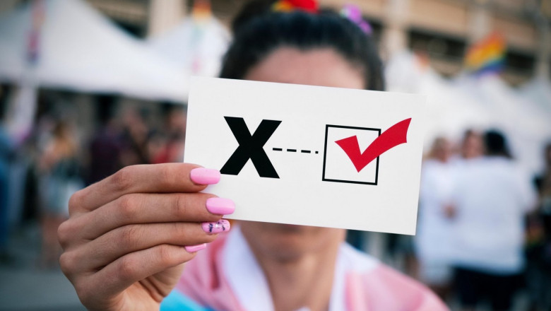 closeup of a young person, wrapped in a transgender pride flag, holding a piece of paper with a check mark on an X letter for the third gender category