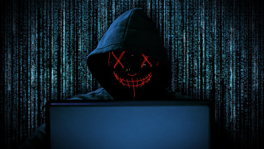 hacker with red glowing mask behind notebook laptop in front of blue source binary code background internet cyber hack attack computer concept