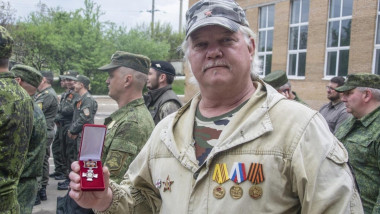 Bărbat cu părul alb și șapcă arată o medalie într-o cutiuță roșie