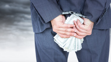 Close up of business man's hands behind back with money and handcuffs against white wall