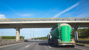 Green cargo tanker truck goes on asphalt highway under concrete bridge, rear view