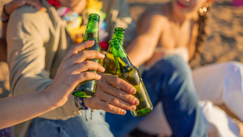 closeup three friends raise their green beer bottles in a toast against the backdrop of a stunning beach sunset. Their camaraderie shines through as t